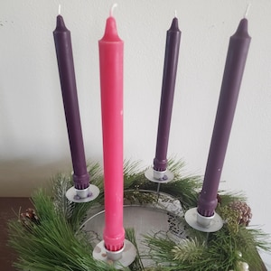 Puede incluir: Una corona de Adviento tradicional con cuatro velas, tres moradas y una rosa, sobre una base de metal con una guirnalda de pino verde y piñas.