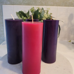 May include: Three pillar candles in shades of purple and red. The candles are cylindrical with a wick at the top. The center candle is a vibrant red, flanked by two deep purple candles. Greenery is visible behind the candles.
