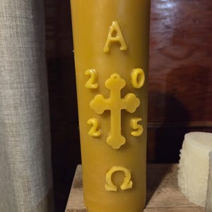 May include: A tall, yellow candle with embossed religious symbols. The candle features the letters "A" and "Ω", the numbers "2025", and a cross. The candle is cylindrical and stands on a wooden surface.