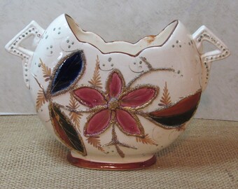 May include: Cream-colored ceramic vase with a wide opening and two handles. The vase is decorated with a large pink flower, blue and black leaves, and gold glitter accents. The base is a dark brown color.