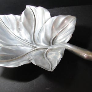 May include: A silver-toned metal dish shaped like a large maple leaf. The dish features a smooth, reflective surface with embossed veins radiating from a central stem that extends outward to form a handle on the right side of the object.