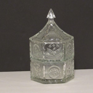 May include: A clear glass candy dish with a lid. The dish is hexagonal and has a design of stars and an eagle on each side. The lid is shaped like a dome.