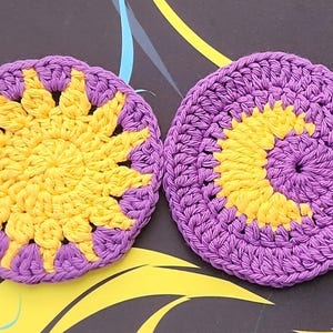 May include: Two handmade crochet coasters. Each coaster features a sun design in yellow and purple. The coasters are circular and made of yarn. The sun design is in the center of each coaster.