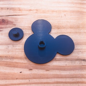 May include: A black plastic Mickey Mouse head shape with two holes in the center. The head is on a wooden surface.