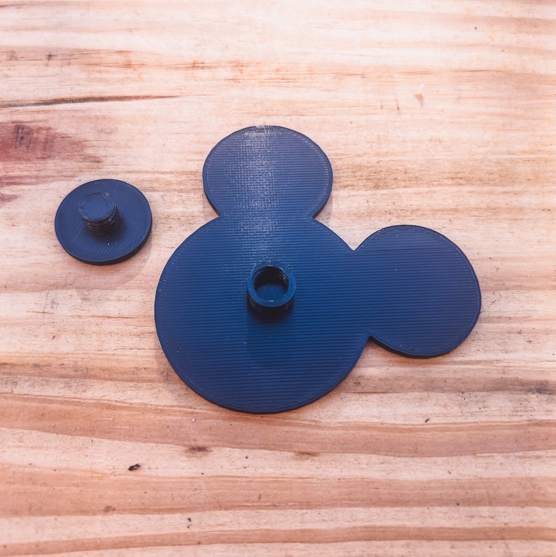 May include: A black plastic Mickey Mouse head shape with two holes in the center. The head is on a wooden surface.