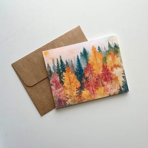 May include: A watercolor painting of a forest scene with trees in shades of red, orange, yellow, and green. The painting is on a card, next to a brown envelope. The card features a landscape of autumn trees.