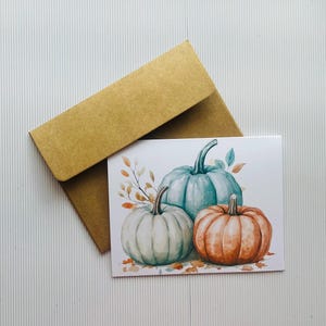 May include: A card with a watercolor painting of three pumpkins. The pumpkins are teal, light green, and orange. The card has a brown envelope.