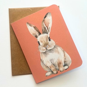 May include: A watercolor illustration of a brown rabbit on a peach-colored background. The rabbit is sitting with its ears up and looking directly at the viewer.