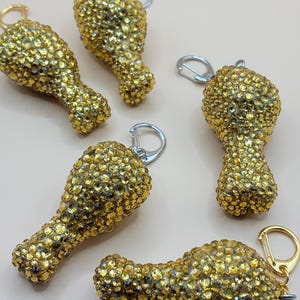 May include: Five gold-colored chicken leg keychains encrusted with sparkling rhinestones. Each keychain has a silver or gold-colored clasp. The keychains are arranged on a light pink surface.