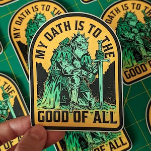 May include: A die-cut sticker featuring a knight in green and black armour kneeling while holding a sword. The background is yellow with the text MY OATH IS TO THE GOOD OF ALL printed in black along the top and bottom edges of the sticker design.