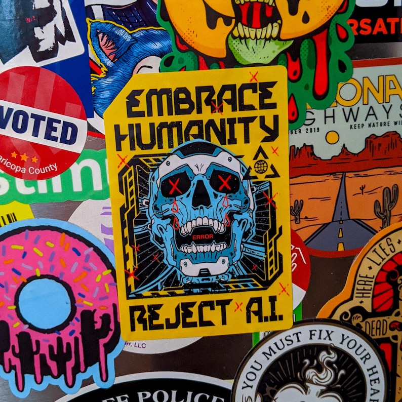Pu&ograve; includere: Una collezione di adesivi con vari design. L'adesivo centrale &egrave; giallo con il testo "EMBRACE HUMANITY REJECT A.I." e presenta un teschio blu con occhi "X" rossi. Altri adesivi includono un adesivo "VOTED", una ciambella e un paesaggio desertico.