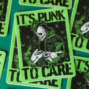 Puede incluir: Pegatina verde con el texto "IT'S PUNK TO CARE" y un gráfico de una figura con cara de calavera con una chaqueta verde sosteniendo un conejo blanco. El diseño tiene una estética punk rock con acentos negros y un fondo radiante.