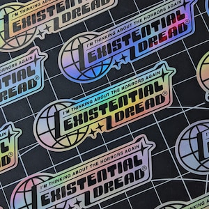 May include: Holographic stickers with the text "I'M THINKING ABOUT THE HORRORS AGAIN" and "EXISTENTIAL DREAD" in a bold, stylized font. The design includes a globe and star accents. The stickers have a rainbow sheen and are on a black grid background.
