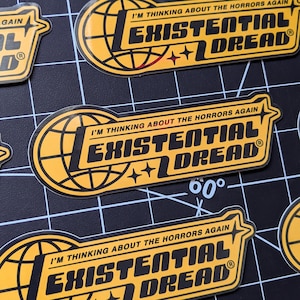 May include: Yellow and black stickers with the text "I'M THINKING ABOUT THE HORRORS AGAIN" and "EXISTENTIAL DREAD". The stickers feature a globe design and star accents. The stickers are on a black grid surface.