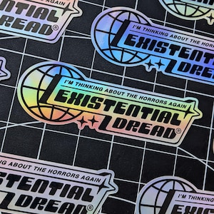 May include: Holographic stickers with the text "I'M THINKING ABOUT THE HORRORS AGAIN EXISTENTIAL DREAD" in a bold, stylized font. The design includes a globe and star accents, with a rainbow sheen. The stickers are on a black grid.