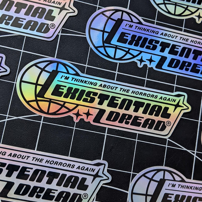 May include: Holographic stickers with the text "I'M THINKING ABOUT THE HORRORS AGAIN EXISTENTIAL DREAD" in a bold, stylized font. The design includes a globe and star accents, with a rainbow sheen. The stickers are on a black grid.