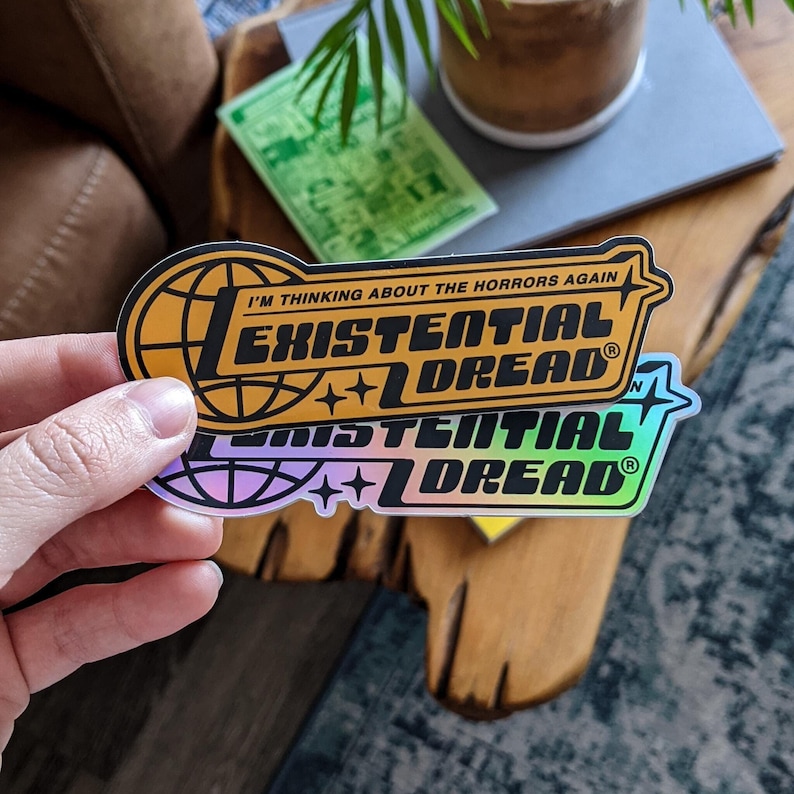 May include: Two stickers with the words "EXISTENTIAL DREAD" in a bold, stylized font. One sticker is orange and black, the other is iridescent. Both stickers have the text "I'M THINKING ABOUT THE HORRORS AGAIN" above the main text.