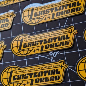 May include: Yellow and black stickers with the text "I'M THINKING ABOUT THE HORRORS AGAIN" and "EXISTENTIAL DREAD". The stickers feature a globe design and star accents. The stickers are on a black grid.