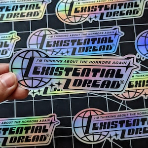 May include: Holographic stickers with the text "I'M THINKING ABOUT THE HORRORS AGAIN" and "EXISTENTIAL DREAD" in a stylized font. The design includes a globe and star accents. The stickers have a reflective, iridescent finish.