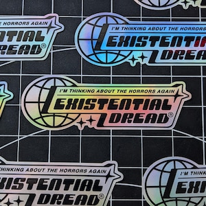 May include: Holographic stickers with the text "I'M THINKING ABOUT THE HORRORS AGAIN" and "EXISTENTIAL DREAD" in a stylized font. The design includes a globe and star accents. The stickers are on a black grid background.