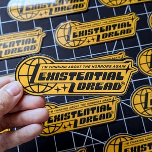 May include: Yellow and black sticker with the text "I'M THINKING ABOUT THE HORRORS AGAIN EXISTENTIAL DREAD". The sticker has a globe and star design. Multiple stickers are visible on a black grid surface.