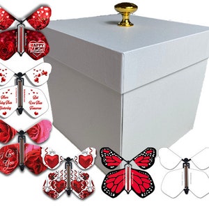 May include: A white gift box with a gold knob on top, surrounded by several butterfly-shaped cards. The cards feature various designs, including roses, hearts, and the text "Happy Valentine's".