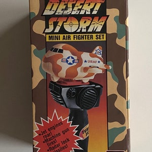 May include: A box for a Desert Storm Mini Air Fighter Set toy. The box is camouflage green and brown with a picture of a toy airplane on the front. The toy airplane is orange, brown, and white with a red cockpit and a black handle. The box also has the text "Electronic Toy Desert Storm Mini Air Fighter Set" and "War in the Gulf" on it.