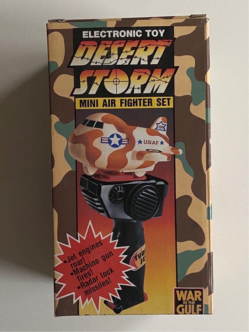 May include: A box for a Desert Storm Mini Air Fighter Set toy. The box is camouflage green and brown with a picture of a toy airplane on the front. The toy airplane is orange, brown, and white with a red cockpit and a black handle. The box also has the text "Electronic Toy Desert Storm Mini Air Fighter Set" and "War in the Gulf" on it.