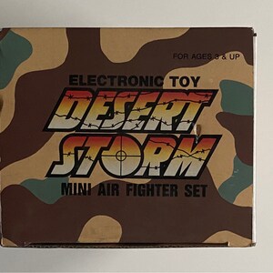 May include: A box for an electronic toy called "Desert Storm Mini Air Fighter Set". The box is brown and tan with a camouflage pattern and the words "Electronic Toy" and "For Ages 3 & Up" printed on it.