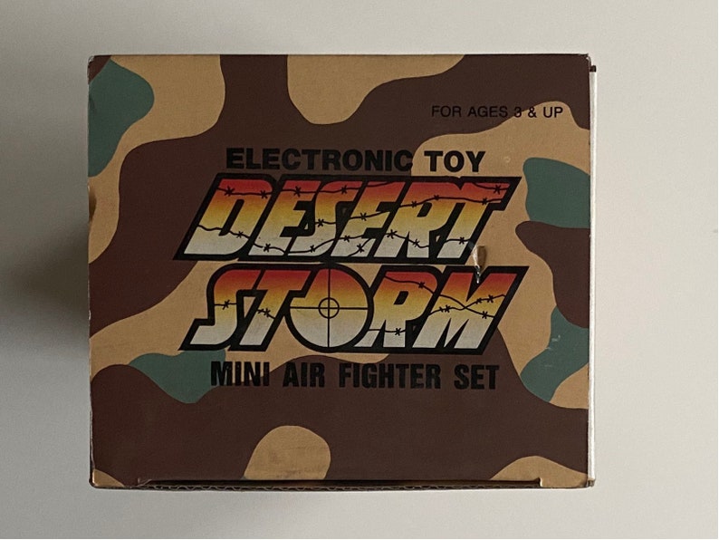 May include: A box for an electronic toy called "Desert Storm Mini Air Fighter Set". The box is brown and tan with a camouflage pattern and the words "Electronic Toy" and "For Ages 3 & Up" printed on it.