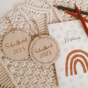 May include: Two wooden ornaments with the text "Schulkind 2023" on them. The ornaments are on a white macrame background. A small notebook with the text "Mein Tier Malbuch" and a rainbow design is in the background.