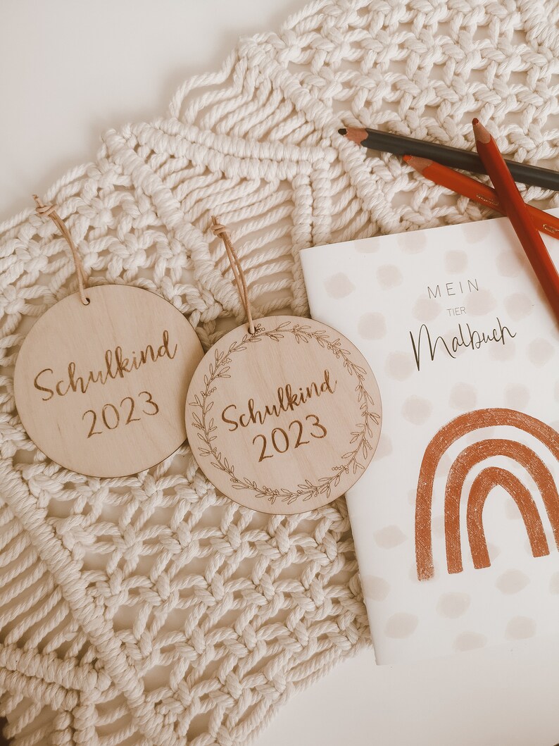 May include: Two wooden ornaments with the text "Schulkind 2023" on them. The ornaments are on a white macrame background. A small notebook with the text "Mein Tier Malbuch" and a rainbow design is in the background.