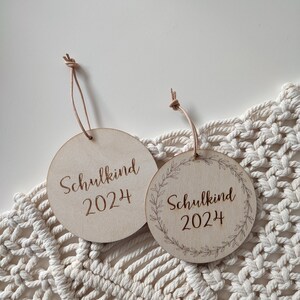 May include: Two round wooden ornaments with the text "Schulkind 2024" engraved on them. One ornament has a simple design, while the other has a wreath design around the text.