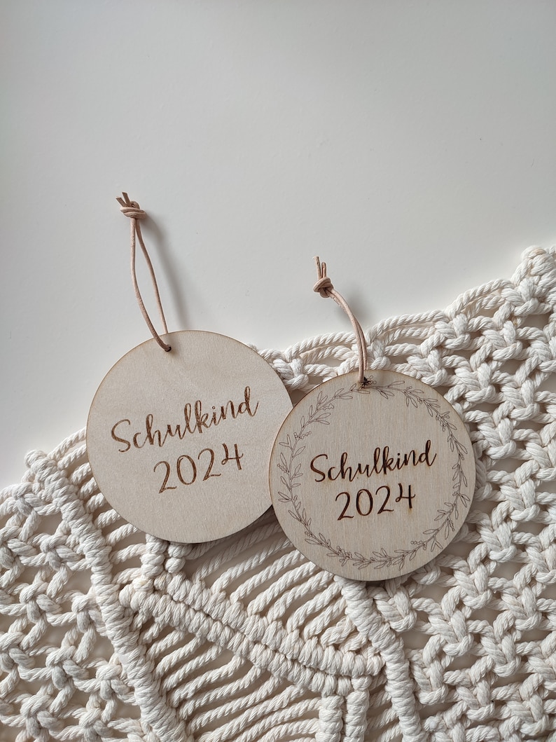 May include: Two round wooden ornaments with the text "Schulkind 2024" engraved on them. One ornament has a simple design, while the other has a wreath design around the text.