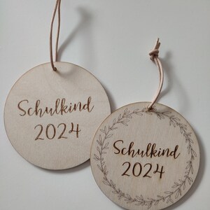 May include: Two round wooden ornaments with the text "Schulkind 2024" on them. One ornament has a simple design and the other has a wreath of leaves around the text.