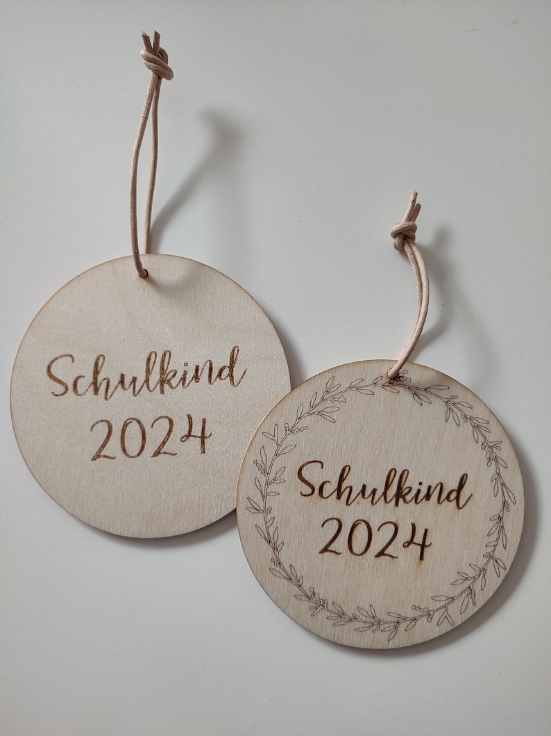 May include: Two round wooden ornaments with the text "Schulkind 2024" on them. One ornament has a simple design and the other has a wreath of leaves around the text.