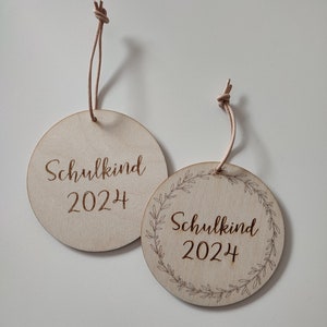 May include: Two round wooden ornaments with the text "Schulkind 2024" engraved on them. One ornament has a simple design, while the other has a wreath of leaves around the text.