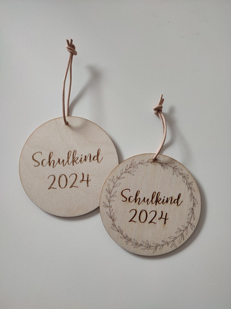May include: Two round wooden ornaments with the text "Schulkind 2024" engraved on them. One ornament has a simple design, while the other has a wreath of leaves around the text.