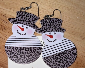 May include: A pair of snowman earrings with a black and white striped pattern and a leopard print design. The earrings are on a white card with the text "Mazie's Charming Treasures".
