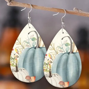 May include: Teardrop-shaped earrings featuring a light blue pumpkin design with autumn foliage. The earrings have silver-colored fish hook ear wires. The background is a cream color.