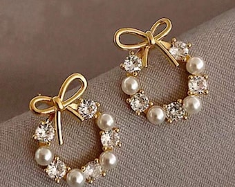 May include: A pair of gold-toned stud earrings. Each earring features a bow at the top and a wreath-like design. The wreath is adorned with faux pearls and clear, sparkling crystals. The earrings are set against a neutral background.