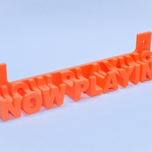 Puede incluir: Un letrero de plástico naranja con las palabras "NOW PLAYING" en letras en relieve. El letrero está diseñado para ser montado en una pared.