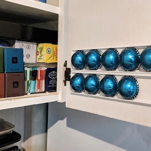 Op de afbeelding: Een witte plastic houder met acht blauwe koffiepads bevestigd aan de binnenkant van een witte keukendeur. De houder is gelabeld "VERTUO" op de dozen met koffiepads.
