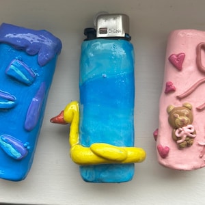 Puede incluir: Una colección de encendedores decorativos. Un encendedor azul presenta un flotador de pato amarillo. Otro encendedor azul está junto a un encendedor rosa decorado con corazones y un osito de peluche. Los encendedores son hechos a mano.