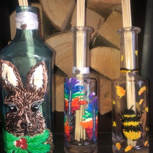 May include: Three decorative glass diffuser bottles with wooden reeds. One bottle features a brown bunny design with a green holly collar. Another has colorful feather art, and the third has a bumblebee design. Each bottle has a ribbon and charm.