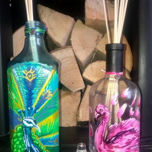 May include: Two glass bottles with painted designs. The bottle on the left has a peacock painted on it with green, blue, and gold colours. The bottle on the right has a pink flamingo painted on it with pink and white colours. Both bottles have wooden sticks inside.