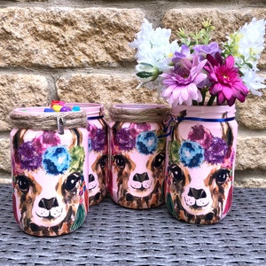 May include: Three pink glass jars with llama illustrations and floral designs. The jars are decorated with brown twine and have a variety of colourful beads inside. The jars are sitting on a grey wicker surface. A bouquet of pink, purple and white flowers is in the background.