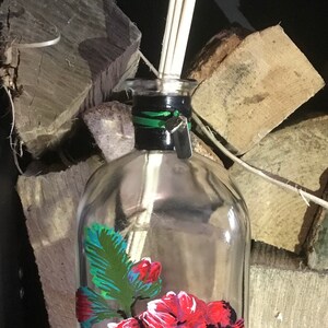 May include: A clear glass diffuser bottle with a black and green neck, decorated with a vibrant red floral design. The bottle contains wooden reeds for fragrance diffusion. The background features stacked firewood.