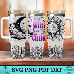 May include: A sublimation design for a tumbler featuring a black and white illustration of a crescent moon, sun, and crystals. The text "Stay Wild Moon Child" is written in purple on the middle tumbler.