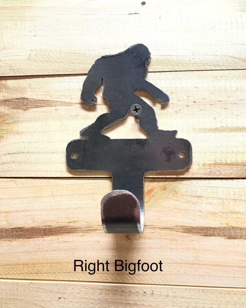 Op de afbeelding: Een metalen wandhaak in de vorm van een Bigfoot-silhouet. De haak is zilverkleurig en heeft een enkele haak aan de onderkant. De tekst "Right Bigfoot" is onder de haak afgedrukt.